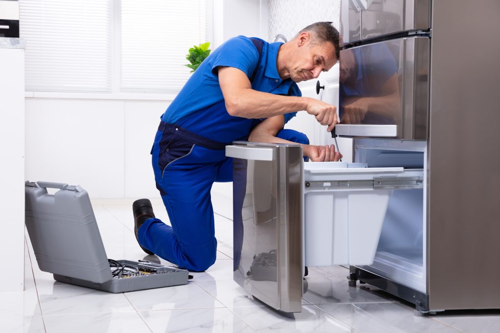 Mature male serviceman providing appliance repair services by fixing a refrigerator in a modern kitchen with a toolbox.